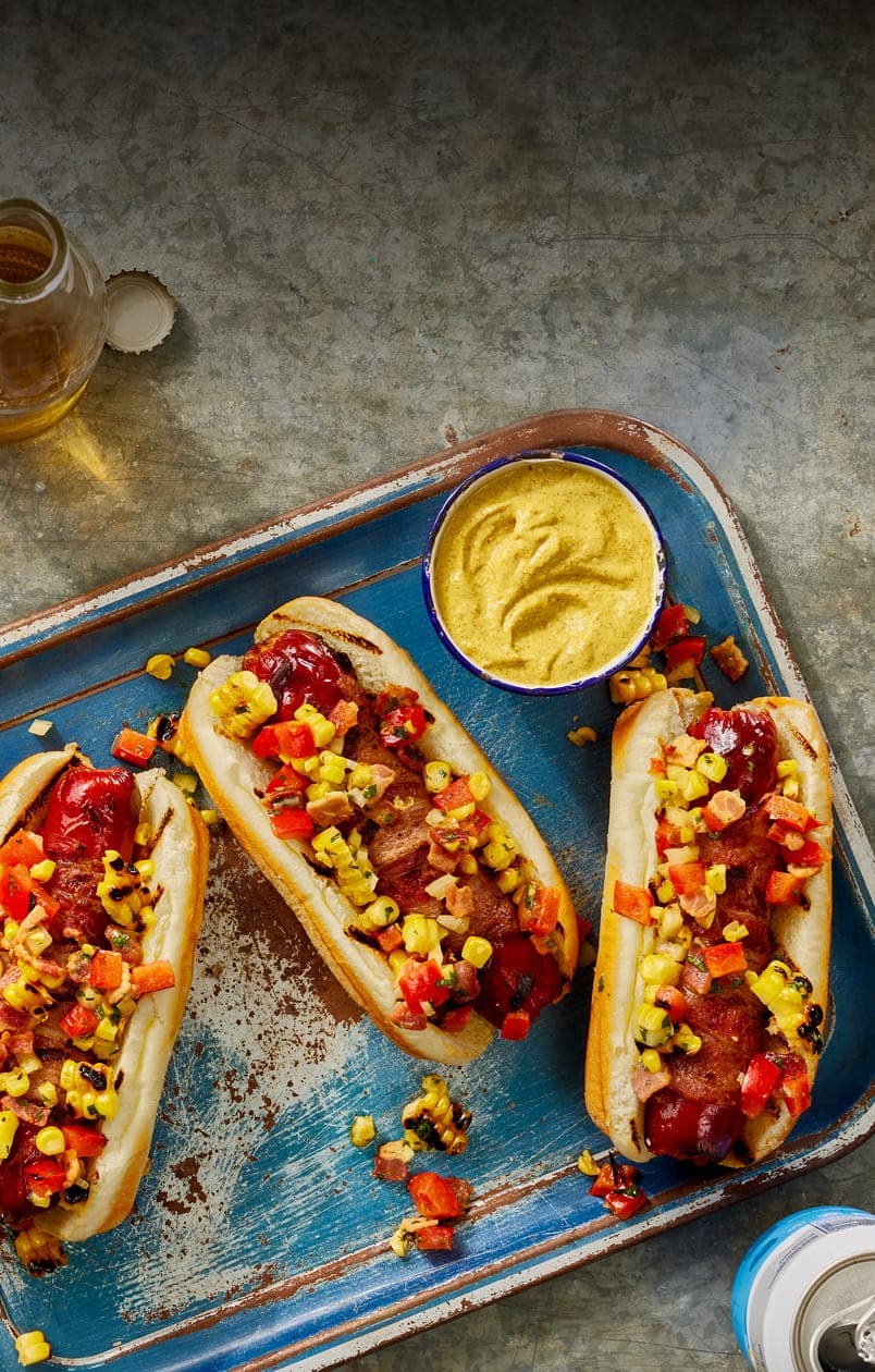 Grilled hot dogs topped with corn salsa and bacon on a tray with mustard dipping sauce.